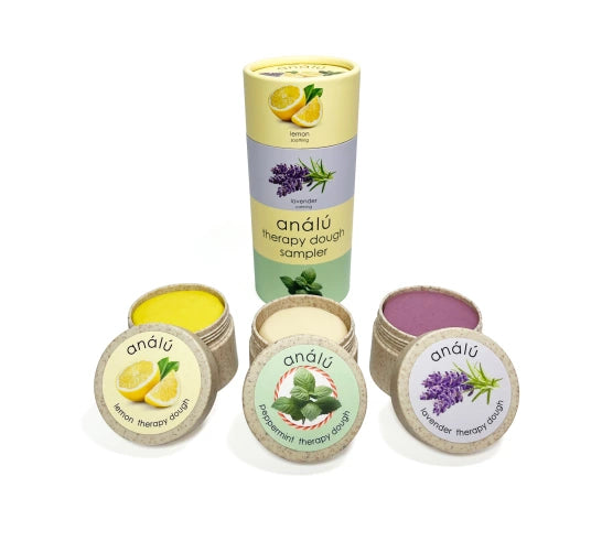 Three open containers of therapy dough—lemon (yellow), peppermint (white), and lavender (purple)—sit in front of a cylindrical box labeled "análú therapy dough sampler." Each container has a matching illustrated lid featuring its scent.