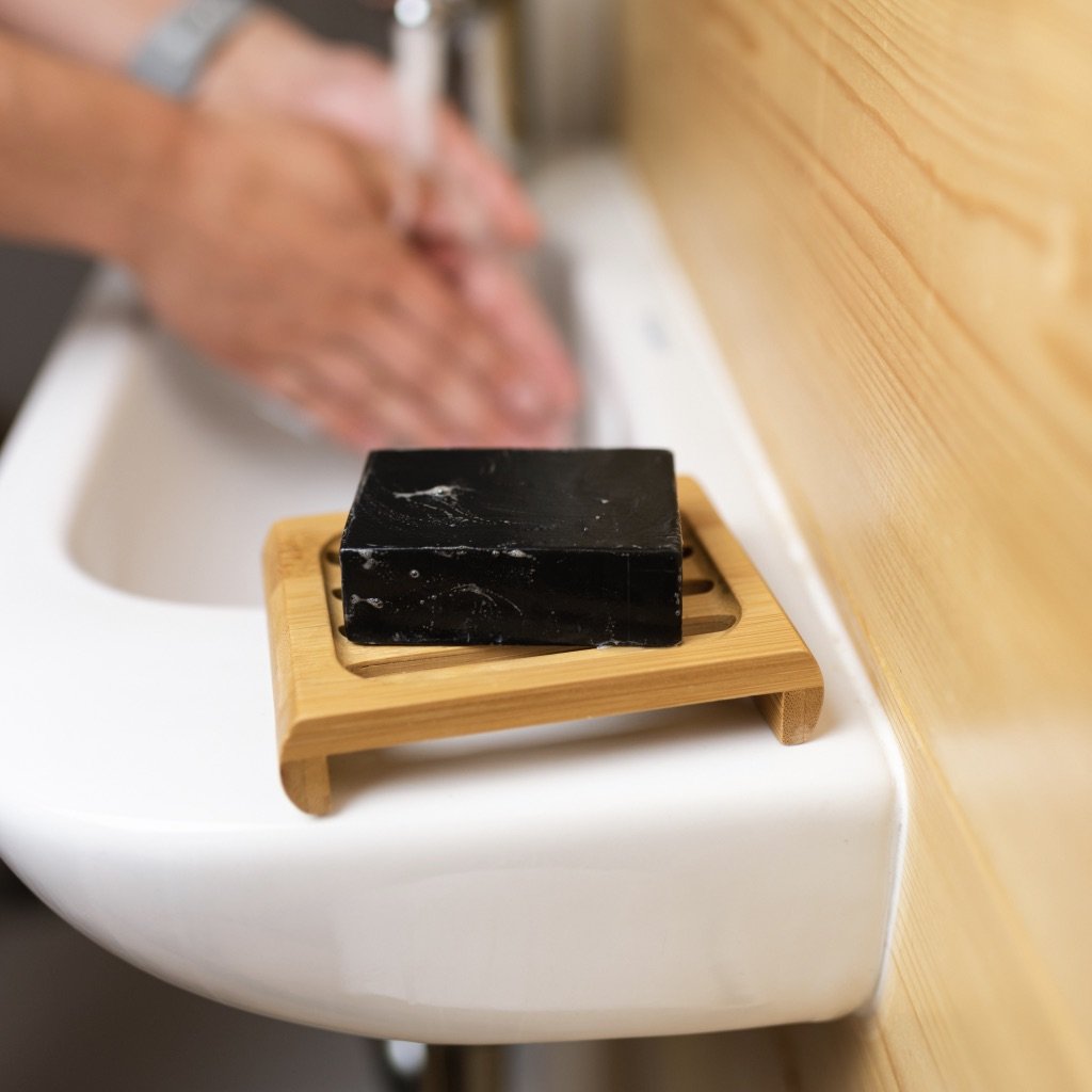 This image showcases a **bamboo soap dish** holding a **black bar of soap** on the edge of a white sink. In the background, a person is washing their hands under running water. The soap dish features a **slatted design**, allowing water to drain efficiently, helping the soap stay dry and last longer. The combination of **eco-friendly bamboo** and the minimalist design makes it a stylish and sustainable bathroom accessory.