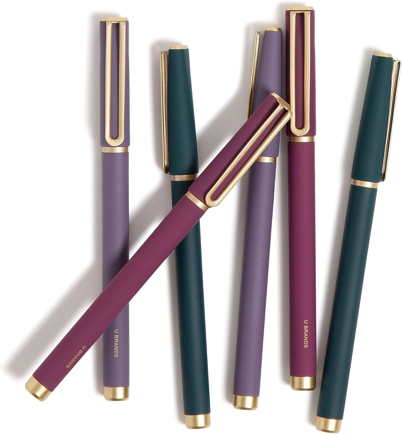 Six pens in rich jewel tones (purple, teal, and burgundy) with matte finishes and gold accents, arranged diagonally on a white surface.