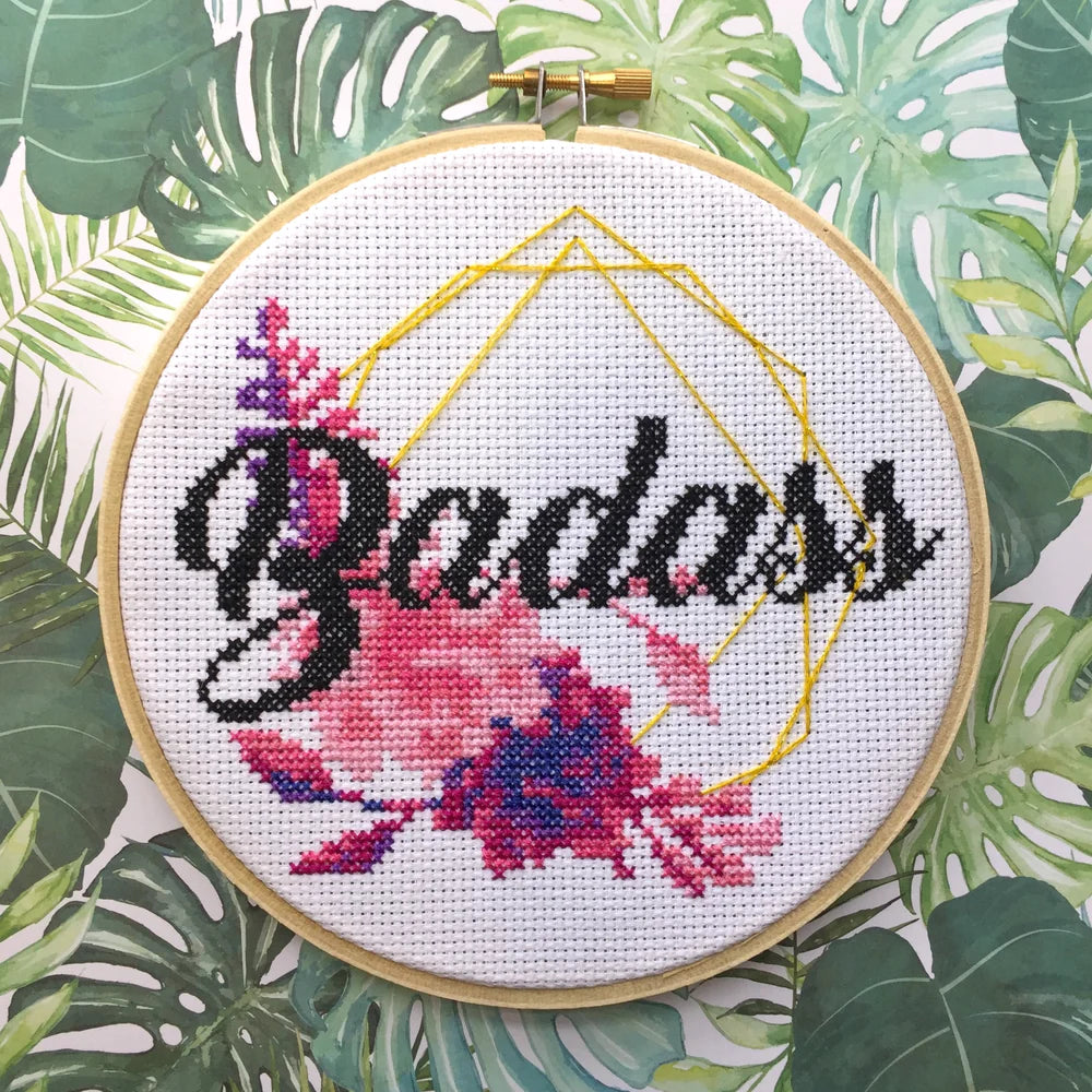 Colorful cross-stitch hoop with the word “Badass” stitched in bold script, surrounded by pink and purple flowers.