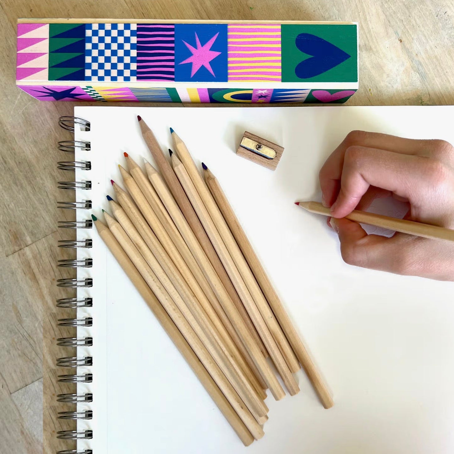 Colored pencil set with playful box design, sharpened pencils on a sketchpad, and a hand drawing on blank paper with a red pencil.