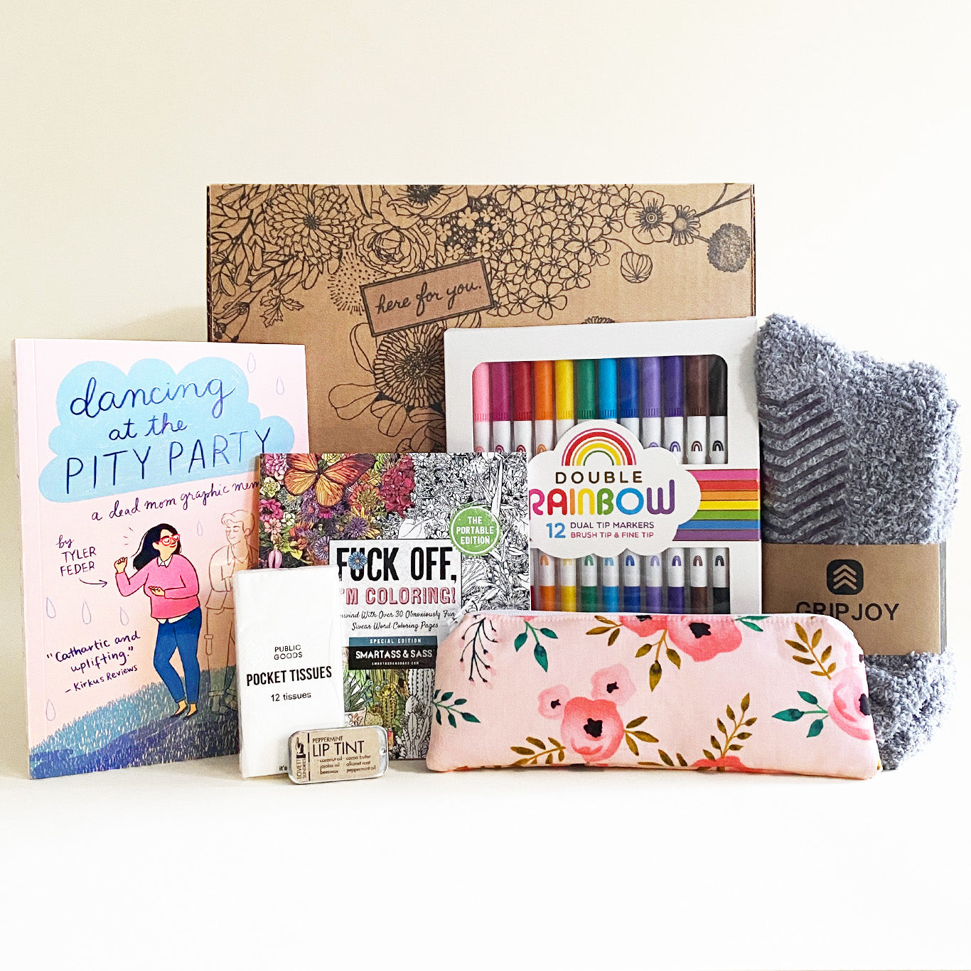 A grief care package from Here For You is displayed against a neutral background. The package includes a cardboard box with a floral design and "here for you" printed on it. Inside are comforting and creative items:
Dancing at the Pity Party by Tyler Feder, a graphic memoir about losing a mother
A portable coloring book titled Fck Off, I’m Coloring!*
A pack of "Double Rainbow" dual-tip markers
A soft, fuzzy GripJoy blanket
A floral pencil case
Pocket tissues
A tube of lip tint
The items are arranged neatly