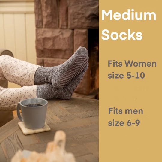 This image is an advertisement for **Medium Socks**, showing a person lounging by a cozy fireplace with their feet up on a wooden table, wearing soft, gray, grippy socks. A warm beverage sits nearby, enhancing the cozy atmosphere. On the right side, the text indicates the sock sizing:
- **Fits Women’s sizes 5-10**
- **Fits Men’s sizes 6-9**
The design and setting emphasize comfort, warmth, and relaxation.