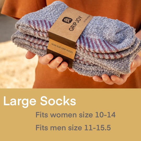 This image is an advertisement for **Large Socks** from Grip Joy. It features a person holding a neatly stacked bundle of soft, gray, non-slip grip socks wrapped in branded packaging. The background has a warm, outdoor setting. On the lower portion of the image, the text highlights the sock sizing:
- **Fits Women’s sizes 10-14**
- **Fits Men’s sizes 11-15.5**
The ad emphasizes comfort, functionality, and a snug fit for larger foot sizes.
