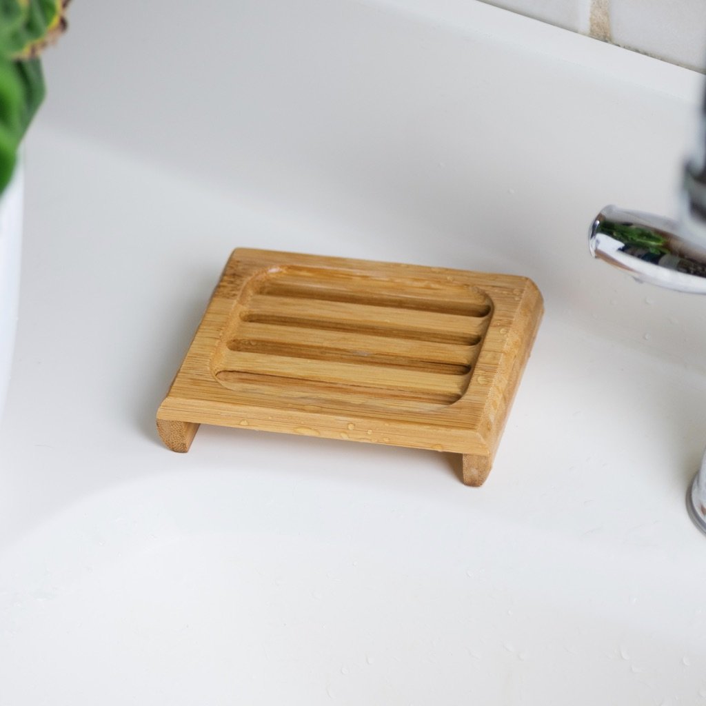 This image features a **bamboo soap dish** placed on a white sink countertop. The dish has a slatted design with raised edges, allowing excess water to drain and air to circulate, keeping soap dry and lasting longer. Its **eco-friendly bamboo construction** makes it a sustainable alternative to plastic soap dishes. With a natural and minimalist aesthetic, it's a great addition to an environmentally conscious bathroom or kitchen setup.