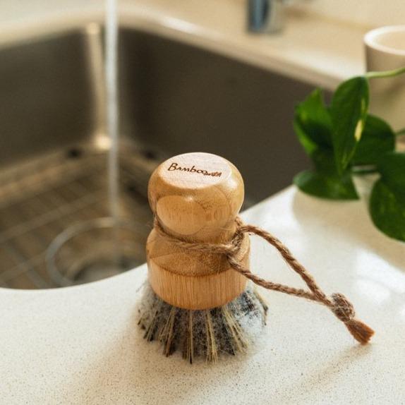 This image features a **bamboo pot scrubber** being used to clean a soapy frying pan. The scrubber has a rounded wooden handle for a comfortable grip and stiff plant-based bristles designed for scrubbing tough residues without damaging surfaces. This eco-friendly kitchen tool is a sustainable alternative to plastic scrubbers, making it an excellent choice for those looking to reduce waste and use biodegradable materials in their home.