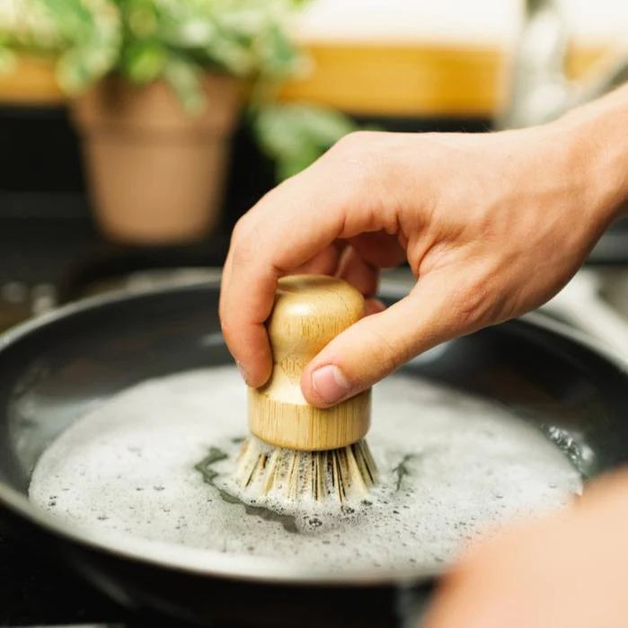 This image features a **bamboo pot scrubber** being used to clean a soapy frying pan. The scrubber has a rounded wooden handle for a comfortable grip and stiff plant-based bristles designed for scrubbing tough residues without damaging surfaces. This eco-friendly kitchen tool is a sustainable alternative to plastic scrubbers, making it an excellent choice for those looking to reduce waste and use biodegradable materials in their home.