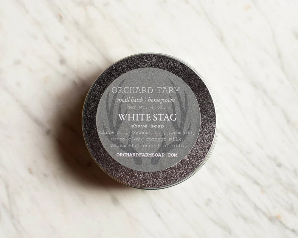 This image features a **tin of "White Stag" shave soap** from Orchard Farm. This **handcrafted, small-batch shaving soap** is made with **olive oil, coconut oil, palm oil, green clay, coconut milk, and balsam-fir essential oils**, creating a nourishing lather for a smooth, irritation-free shave. The **natural ingredients** provide hydration and protection, making it an ideal choice for those seeking a **gentle, botanical-based grooming experience.**