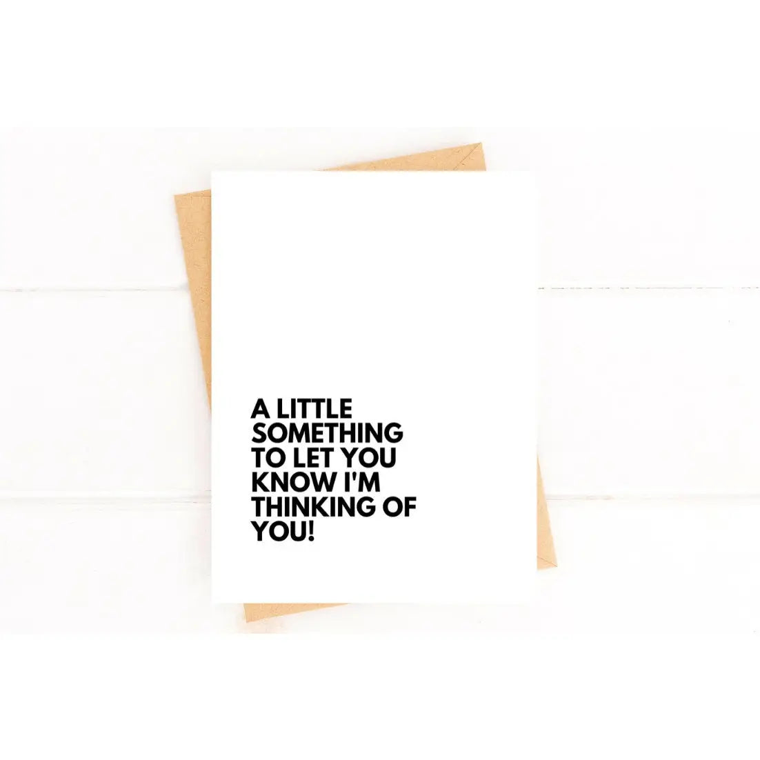 This image features a **minimalist greeting card** with a heartfelt message printed in bold black text:
**"A little something to let you know I'm thinking of you!"**
The card is displayed on a white surface, with a **natural brown envelope** partially visible behind it. The clean and simple design emphasizes the message, making it a thoughtful way to express care and support to someone going through a difficult time. The neutral aesthetic makes it suitable for various occasions, including **grief, enc