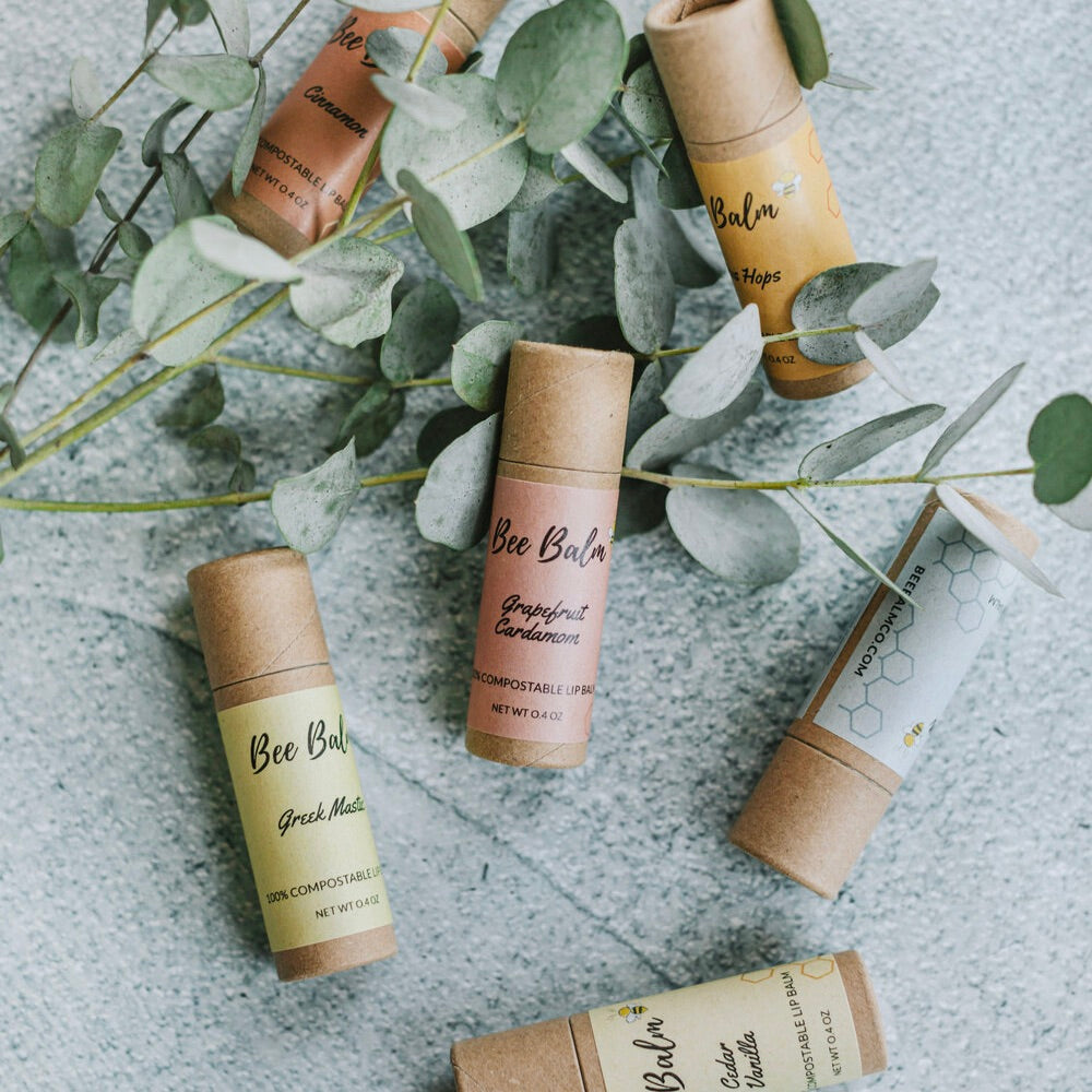 This image showcases **Bee Balm compostable lip balms** in various flavors, including **Grapefruit Cardamom, Greek Mastiha, Cedar Vanilla, Cinnamon, and Hops**. Each tube is made from **eco-friendly, plastic-free packaging**, emphasizing sustainability. The balms are **nourishing and handcrafted**, designed to keep lips soft while supporting **pollinators and the environment**. The natural aesthetic highlights their commitment to **clean, responsible beauty**.