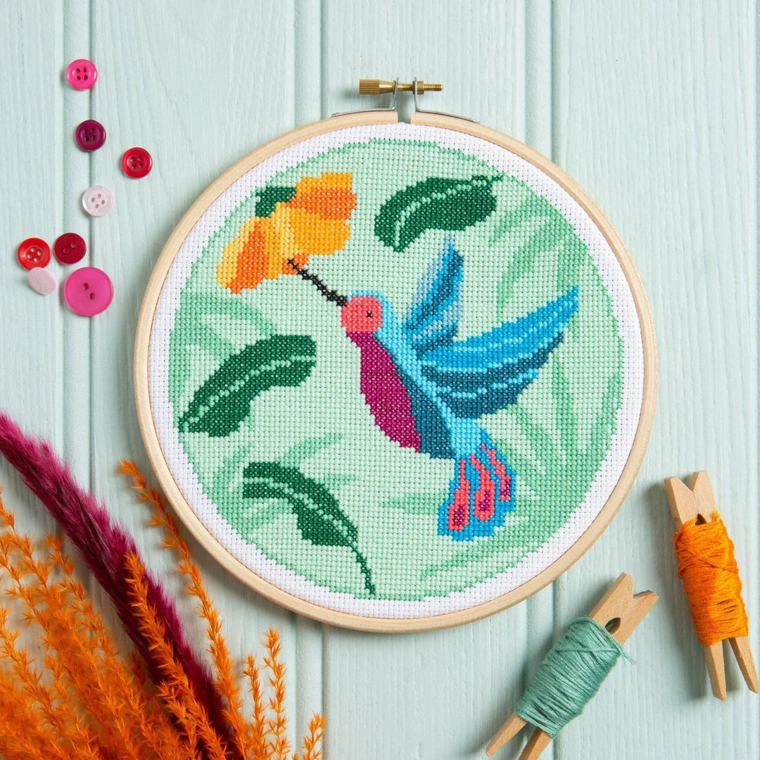 Hummingbird cross-stitch hovering near a yellow flower on green background, with buttons and thread nearby.