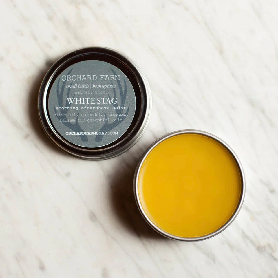 This image showcases a **tin of "White Stag" aftershave salve** from Orchard Farm. Made in **small batches**, this **soothing balm** contains **olive oil, calendula, beeswax, and balsam-fir essential oils** to help calm and moisturize the skin post-shave. The rich **golden salve** is designed to **nourish and protect**, making it a great addition to a natural grooming routine. Perfect for those seeking **botanical, chemical-free skincare.**