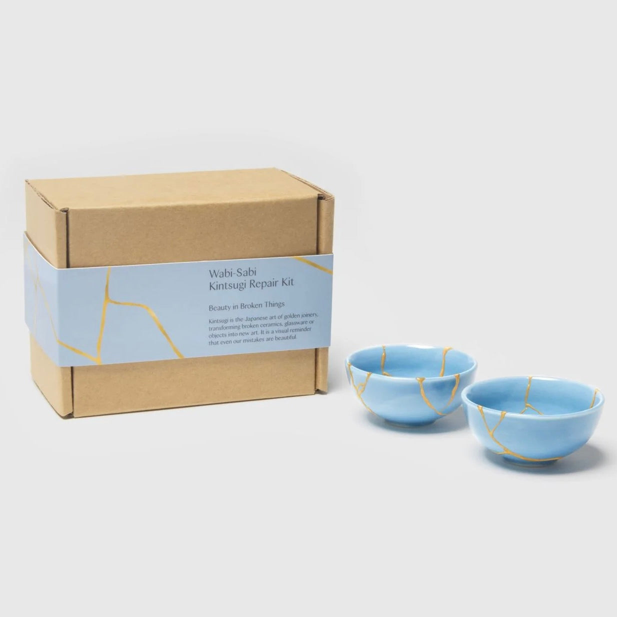 Two blue ceramic bowls with gold repair lines next to a cardboard box labeled 'Wabi Sabi Kintsugi Repair Kit'.