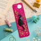 A bright pink fabric bookmark featuring a black cat embroidered in the center, surrounded by green and white floral details and a white sunburst. The bookmark is styled in a flat-lay arrangement with craft props including thread spools, a wooden pencil, paper pom-poms, and an open book.