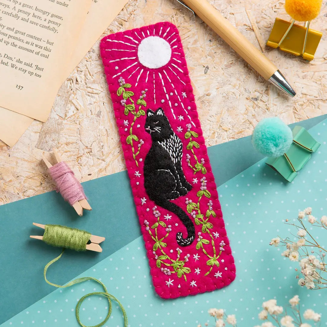A bright pink fabric bookmark featuring a black cat embroidered in the center, surrounded by green and white floral details and a white sunburst. The bookmark is styled in a flat-lay arrangement with craft props including thread spools, a wooden pencil, paper pom-poms, and an open book.