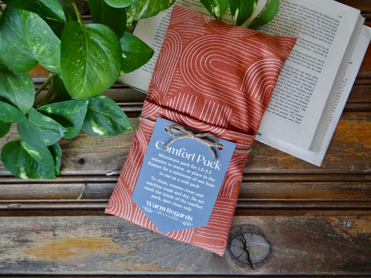 A rust-colored comfort pack with a modern white pattern rests on wood near a plant and open book.