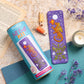 A felt craft kit for making a dragon bookends set, shown with its blue cylindrical packaging and the completed purple bookend featuring an embroidered orange dragon, green leaves, and white flowers. The scene is styled with a book, wooden spools of thread, a lit candle, and dried purple flowers on a patterned teal and wood background.