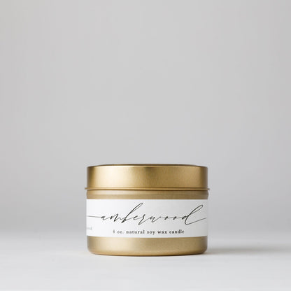 Gold tin candle labeled “amberwood” with a minimalist white label and script font; 4 oz. natural soy wax candle on a light background.