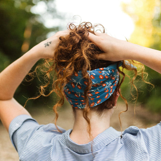 This image captures the back of a woman with curly, auburn hair, secured with a **blue floral-patterned headband**. She is wearing a light blue striped shirt, and her hands are adjusting her hair. A small tattoo with the letters "hv" is visible on her wrist. The background features a blurred natural setting with lush greenery and warm sunlight, giving the photo a **carefree, rustic, and outdoor adventure feel**.
