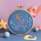 An embroidery hoop features a howling wolf stitched with colorful thread under a golden crescent moon and stars. Surrounded by trees, cosmic details, and whimsical decor on a purple and blue background.