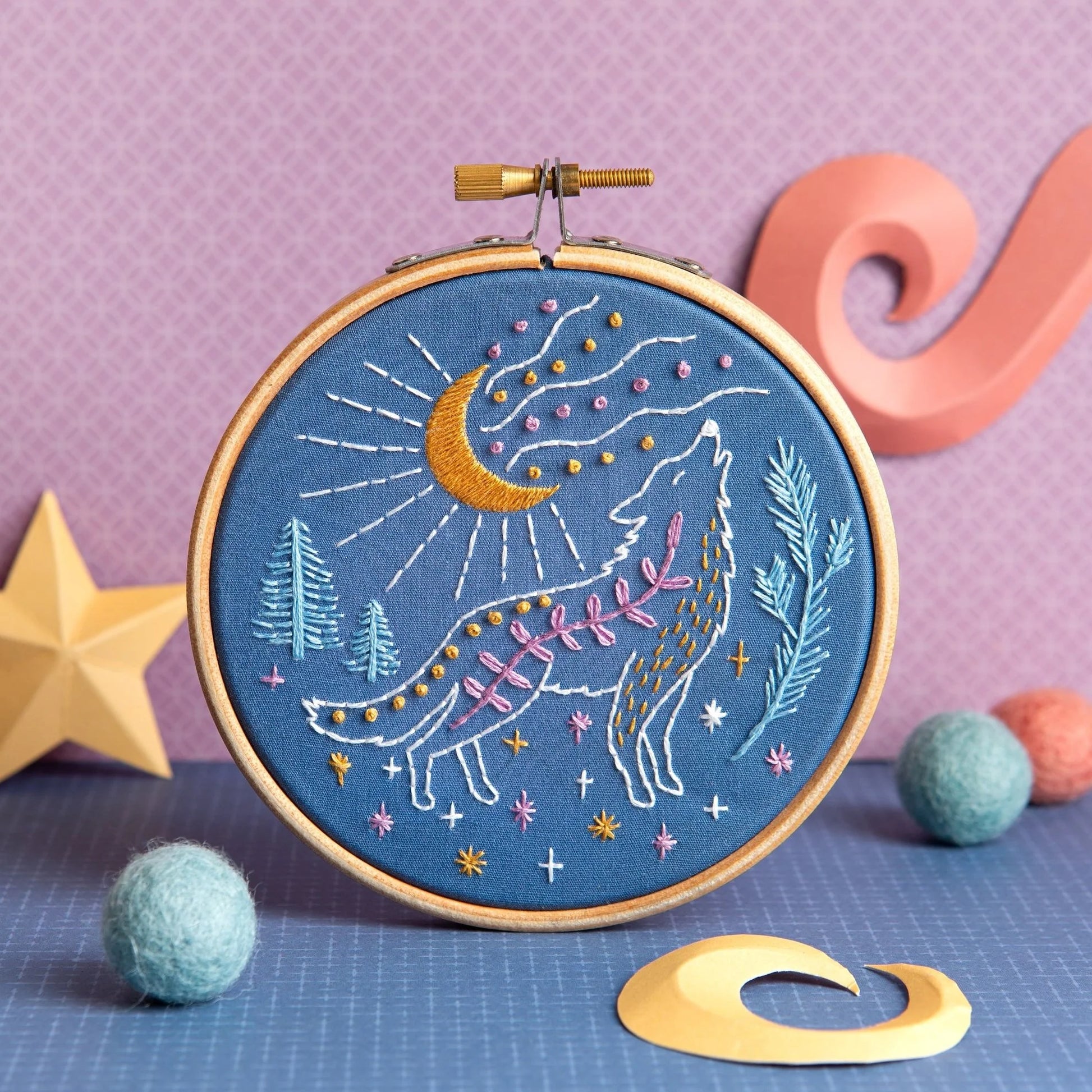 An embroidery hoop features a howling wolf stitched with colorful thread under a golden crescent moon and stars. Surrounded by trees, cosmic details, and whimsical decor on a purple and blue background.