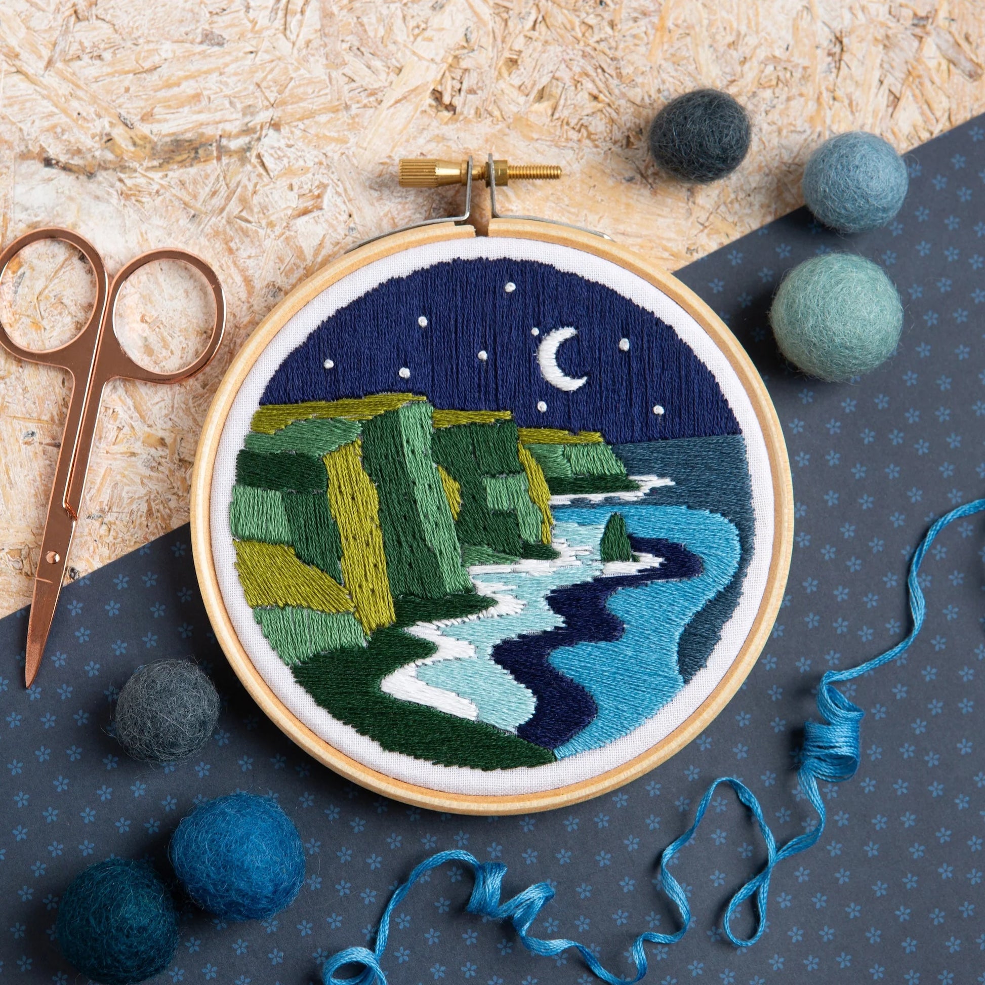 Embroidery hoop with a night scene of cliffs and moonlight, surrounded by embroidery supplies on a textured surface.