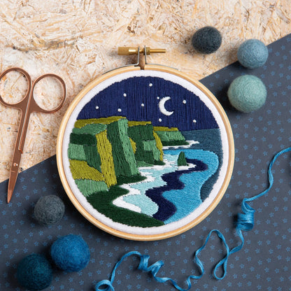 Embroidery hoop with a night scene of cliffs and moonlight, surrounded by embroidery supplies on a textured surface.