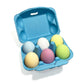 Six colorful egg-shaped bath fizzies in pastel shades sit inside a bright blue egg carton, ready to drop into the tub for fizzy, fun bath time.