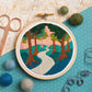An embroidery hoop shows a forest scene with a winding path, tall trees, and a pink sunset sky. Surrounded by felt balls, thread, scissors, and blue polka-dot fabric on a textured surface.
