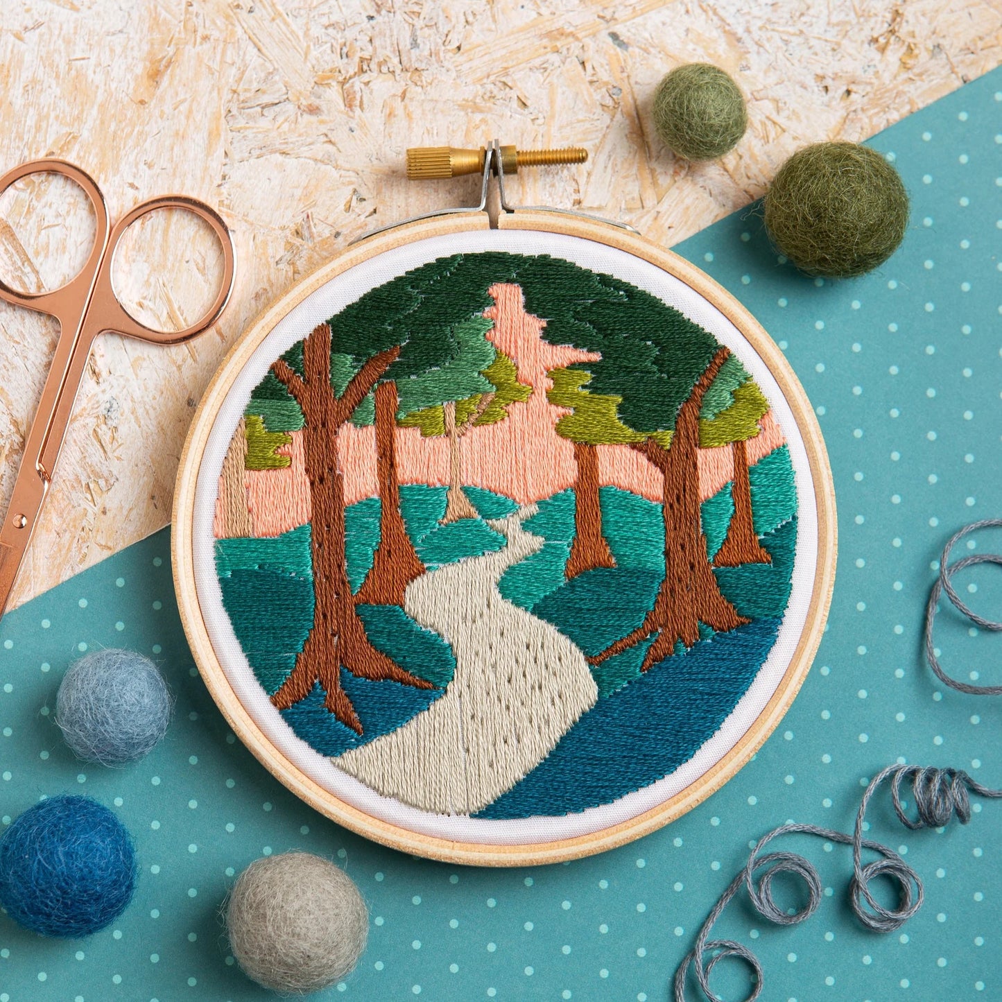 An embroidery hoop shows a forest scene with a winding path, tall trees, and a pink sunset sky. Surrounded by felt balls, thread, scissors, and blue polka-dot fabric on a textured surface.