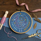 An embroidery hoop displays a colorful, celestial bird stitched on blue fabric with stars, a sun, and a crescent moon. Surrounded by scissors, dried leaves, pink thread, and fabric on a wooden surface.