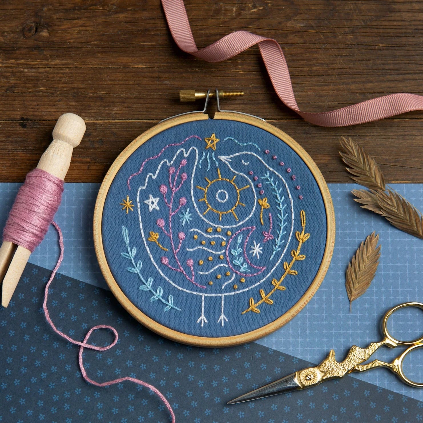 An embroidery hoop displays a colorful, celestial bird stitched on blue fabric with stars, a sun, and a crescent moon. Surrounded by scissors, dried leaves, pink thread, and fabric on a wooden surface.