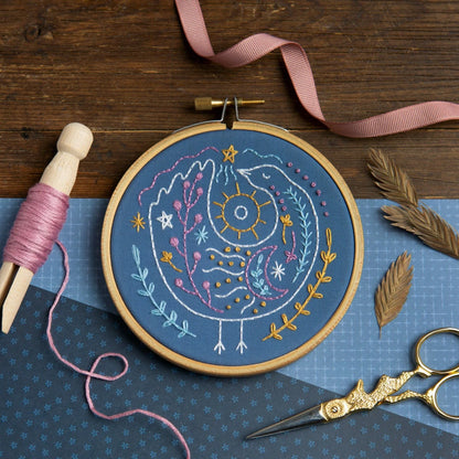 An embroidery hoop displays a colorful, celestial bird stitched on blue fabric with stars, a sun, and a crescent moon. Surrounded by scissors, dried leaves, pink thread, and fabric on a wooden surface.