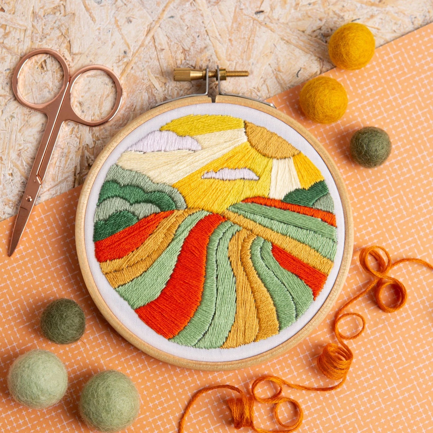 Embroidery hoop with colorful landscape design, surrounded by embroidery tools on a textured surface.
