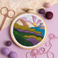 An embroidery hoop features a colorful mountain landscape with layered hills, a lake, and a vibrant sunrise. Surrounded by felt balls, pink thread, and scissors on a textured and patterned background.
