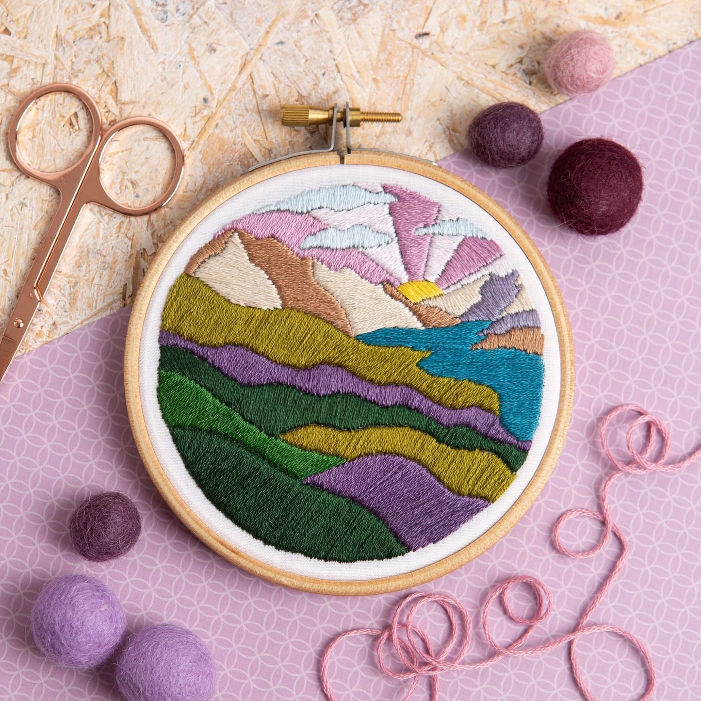 An embroidery hoop features a colorful mountain landscape with layered hills, a lake, and a vibrant sunrise. Surrounded by felt balls, pink thread, and scissors on a textured and patterned background.
