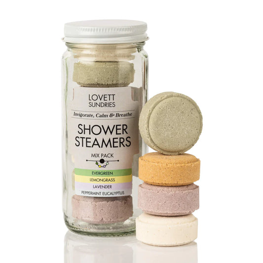 Alt text: A glass jar of Lovett Sundries Shower Steamers is displayed alongside a small stack of individual steamers in different colors. The jar has a white screw-on lid and a label that reads "Invigorate, Calm & Breathe" and "Shower Steamers Mix Pack," listing scents such as Evergreen, Lemongrass, Lavender, and Peppermint Eucalyptus. The steamers are round and slightly textured, with colors including green, orange, purple, and white. One green steamer leans against the stack. The background is plain white