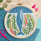 An embroidery hoop features a blue whale surrounded by colorful seaweed and stars on an ocean-blue background. Displayed with thread, scissors, buttons, and patterned papers on a textured surface.