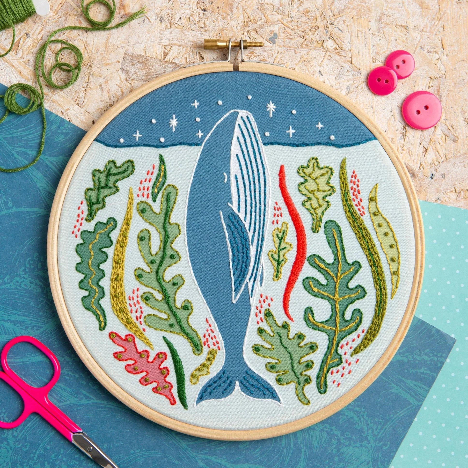 An embroidery hoop features a blue whale surrounded by colorful seaweed and stars on an ocean-blue background. Displayed with thread, scissors, buttons, and patterned papers on a textured surface.