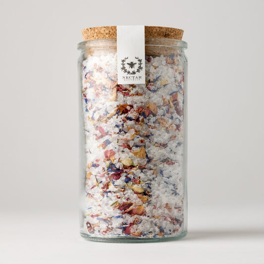 This image features a **glass jar of botanical bath salts** from **Nectar Republic**. The jar is sealed with a **cork lid** and adorned with a minimalist **white label with an elegant floral emblem**. The salts inside are a blend of **coarse sea salt infused with dried flower petals**, including **rose, blue cornflower, and calendula**, creating a visually stunning mix. This luxurious soak is likely designed for **relaxation, skin nourishment, and aromatherapy benefits**.