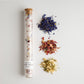 This image showcases a **wildflower-infused bath soak** in a **tall glass test tube** sealed with a **cork stopper**. The soak contains **coarse sea salt blended with dried botanicals**, including **blue cornflower, rose petals, and jasmine flowers**, arranged beside the tube. The label reads **"Apothecary Bath Soak – Wildflowers"**, emphasizing a **natural, soothing, and aromatic** bathing experience. Ideal for **relaxation, skincare, and self-care rituals**.