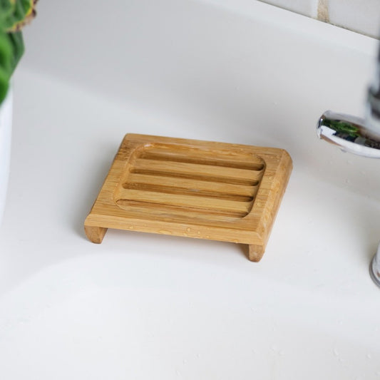 This image features a **bamboo soap dish** placed on a white sink countertop. The dish has a slatted design with raised edges, allowing excess water to drain and air to circulate, keeping soap dry and lasting longer. Its **eco-friendly bamboo construction** makes it a sustainable alternative to plastic soap dishes. With a natural and minimalist aesthetic, it's a great addition to an environmentally conscious bathroom or kitchen setup.