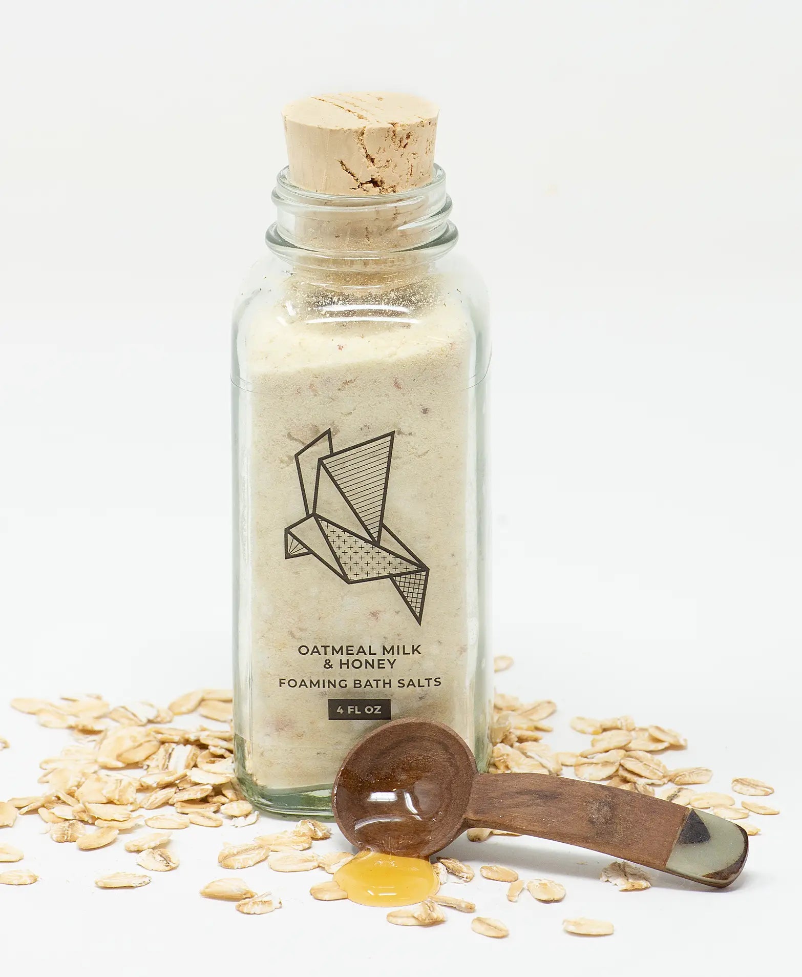 Bottle of oatmeal milk and honey foaming bath salts with a wooden scoop and oats on a white background