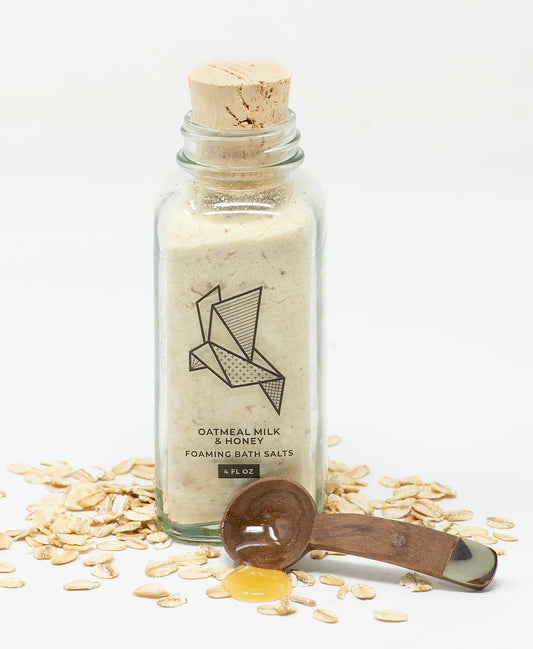 Bottle of oatmeal milk and honey foaming bath salts with a wooden scoop and oats on a white background