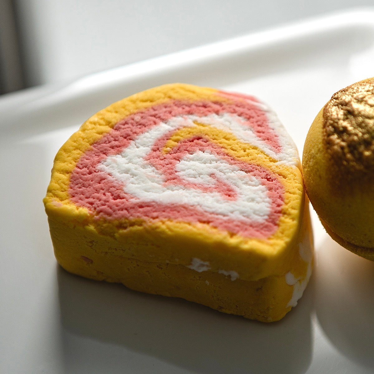 yellow bubble bar on white porcelain surface next to yellow and gold bath bomb. Bubble bar has white and red swirls. 