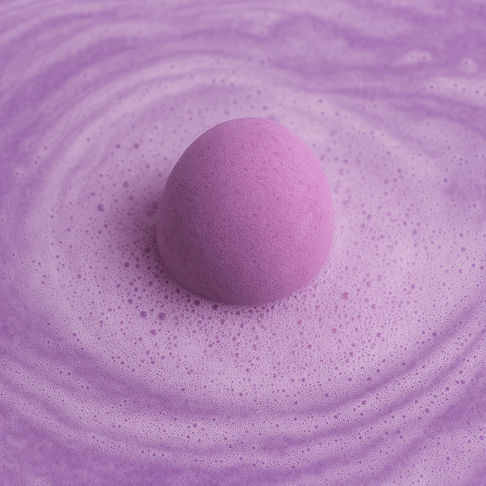 A round, purple bath fizzie floats in bubbly lavender-colored water, creating swirling foam as it dissolves.