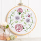 An embroidery hoop displays a garden scene with red roses, green vines, purple flowers, and a bee on white fabric. Decorated with a pink rose in a glass jar and green twine on a light background.
