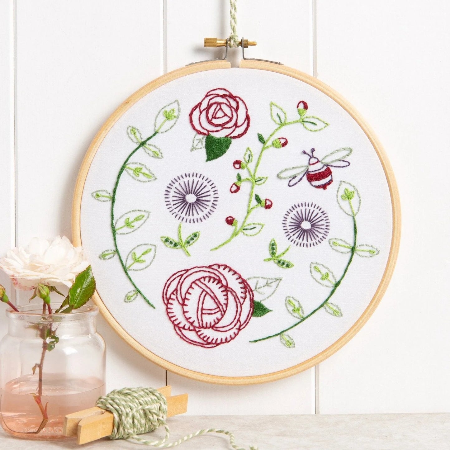 An embroidery hoop displays a garden scene with red roses, green vines, purple flowers, and a bee on white fabric. Decorated with a pink rose in a glass jar and green twine on a light background.

