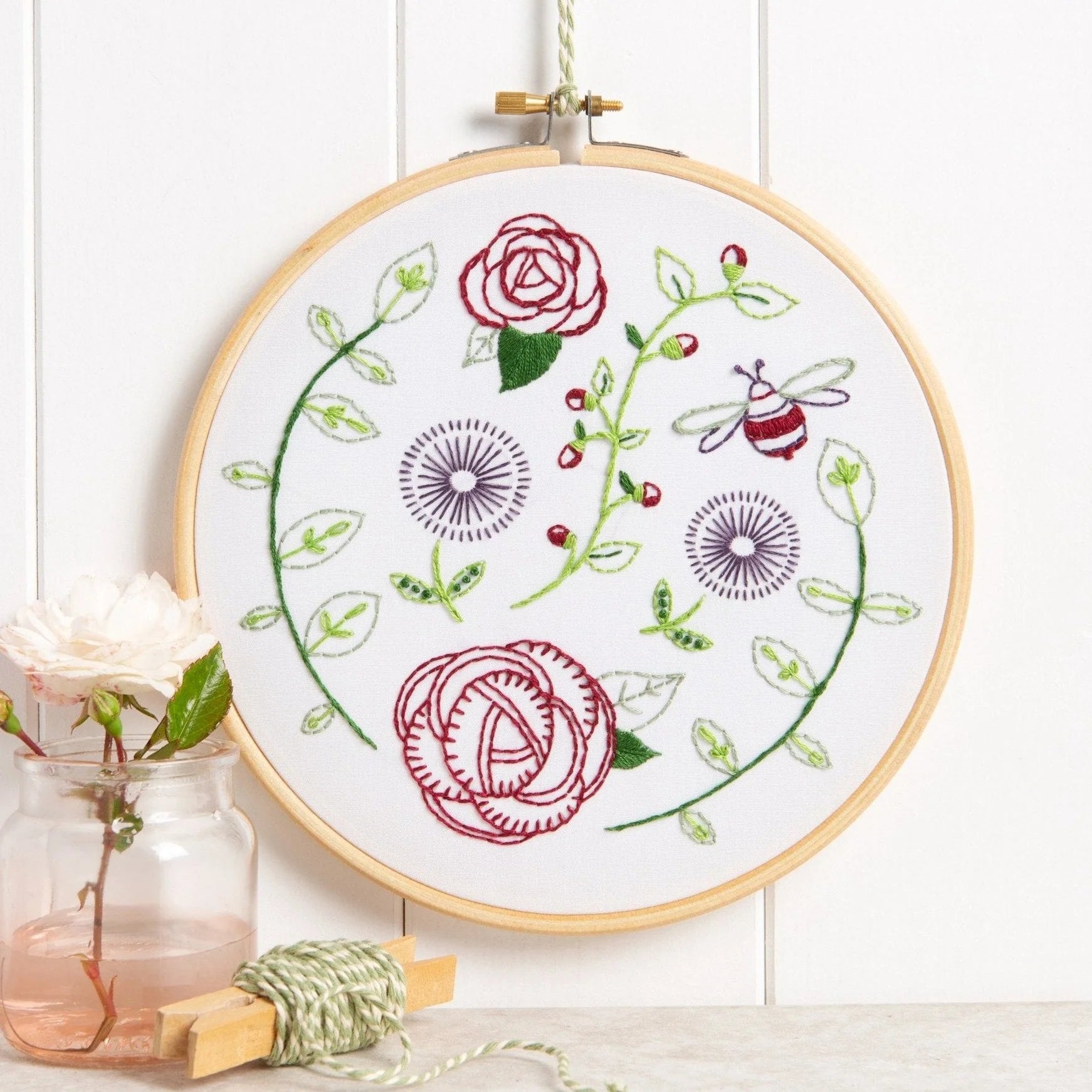 An embroidery hoop displays a garden scene with red roses, green vines, purple flowers, and a bee on white fabric. Decorated with a pink rose in a glass jar and green twine on a light background.
