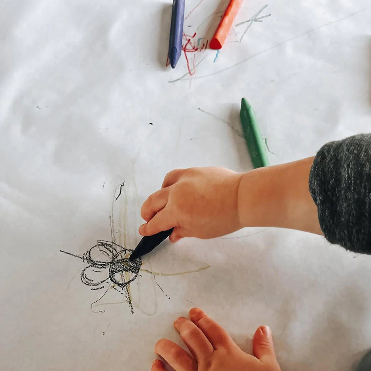 A young child uses a black crayon to draw on white paper, surrounded by colorful triangular crayons in red, green, and blue.